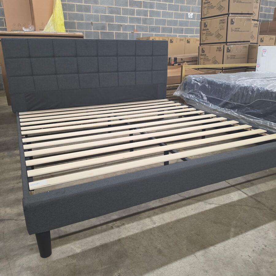 A charcoal upholstered platform bed frame with a square-tufted headboard and wooden slats, in the warehouse.