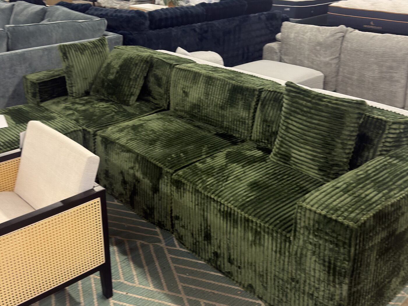 An olive green corduroy modular sectional on a sage herringbone rug, with a cane and black-framed accent chair beside it.