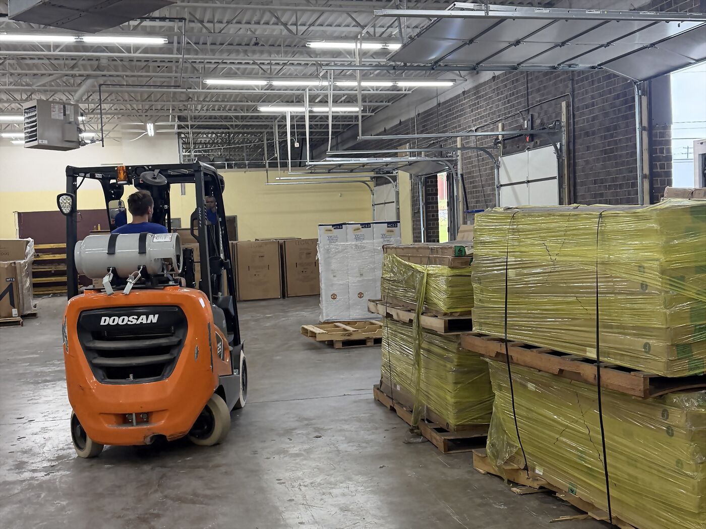 A forklift beside pallets of strapped stock in the warehouse, with a loading door behind.