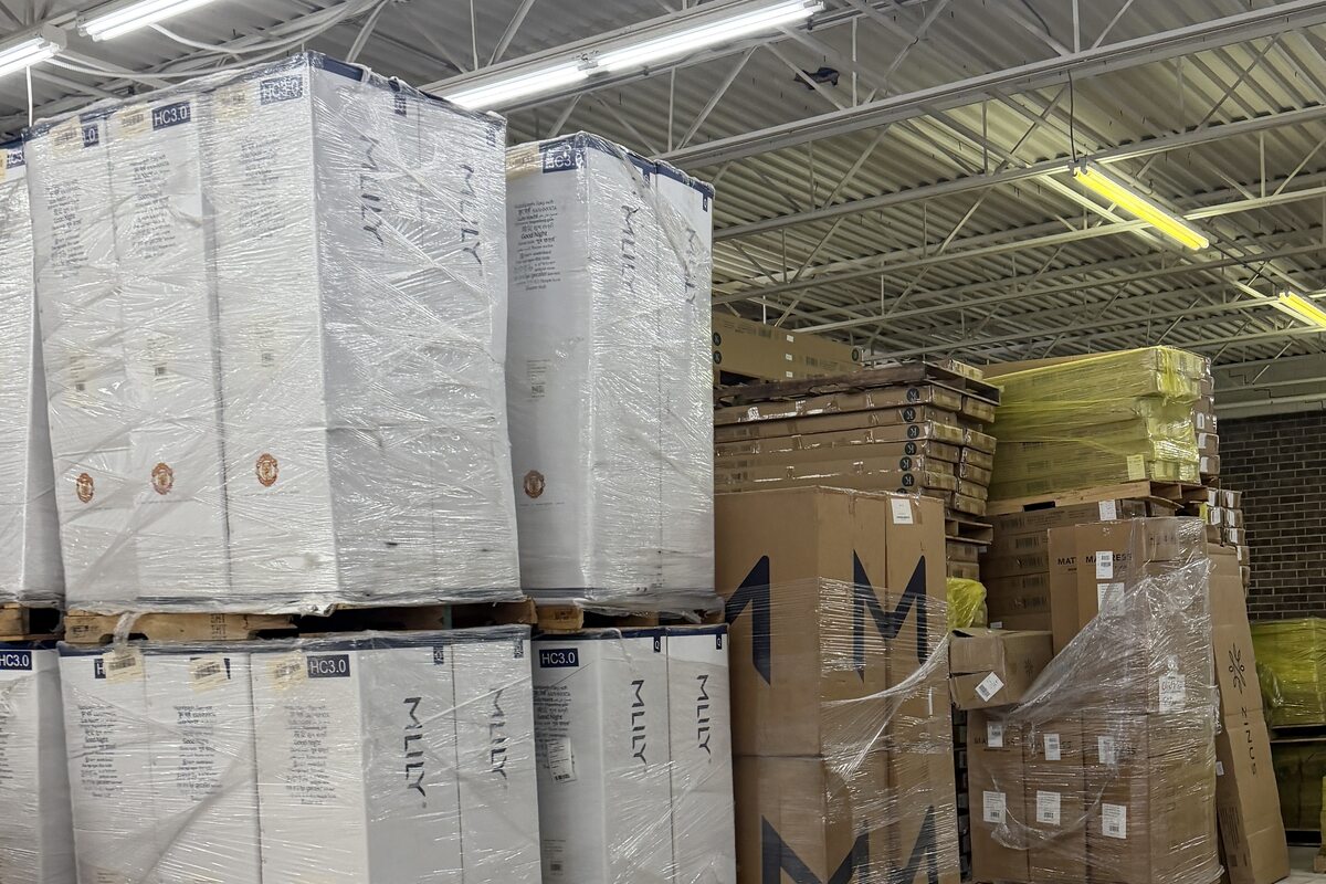 Shrink-wrapped mattresses stacked two pallets high beside cartons of frames and bases.